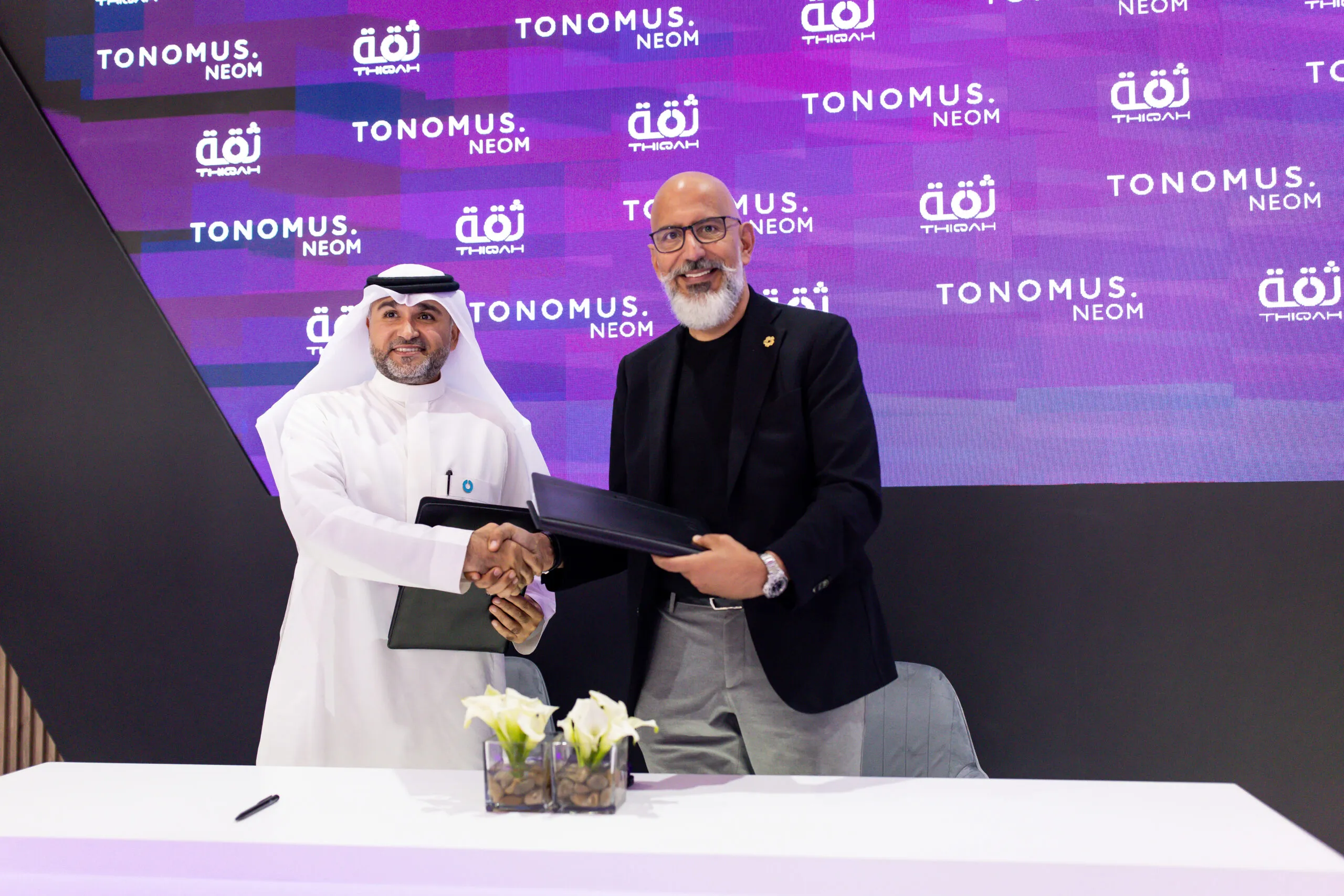 Two executives shaking hands over signed agreements, before a Tonomus and Thiqah backdrop