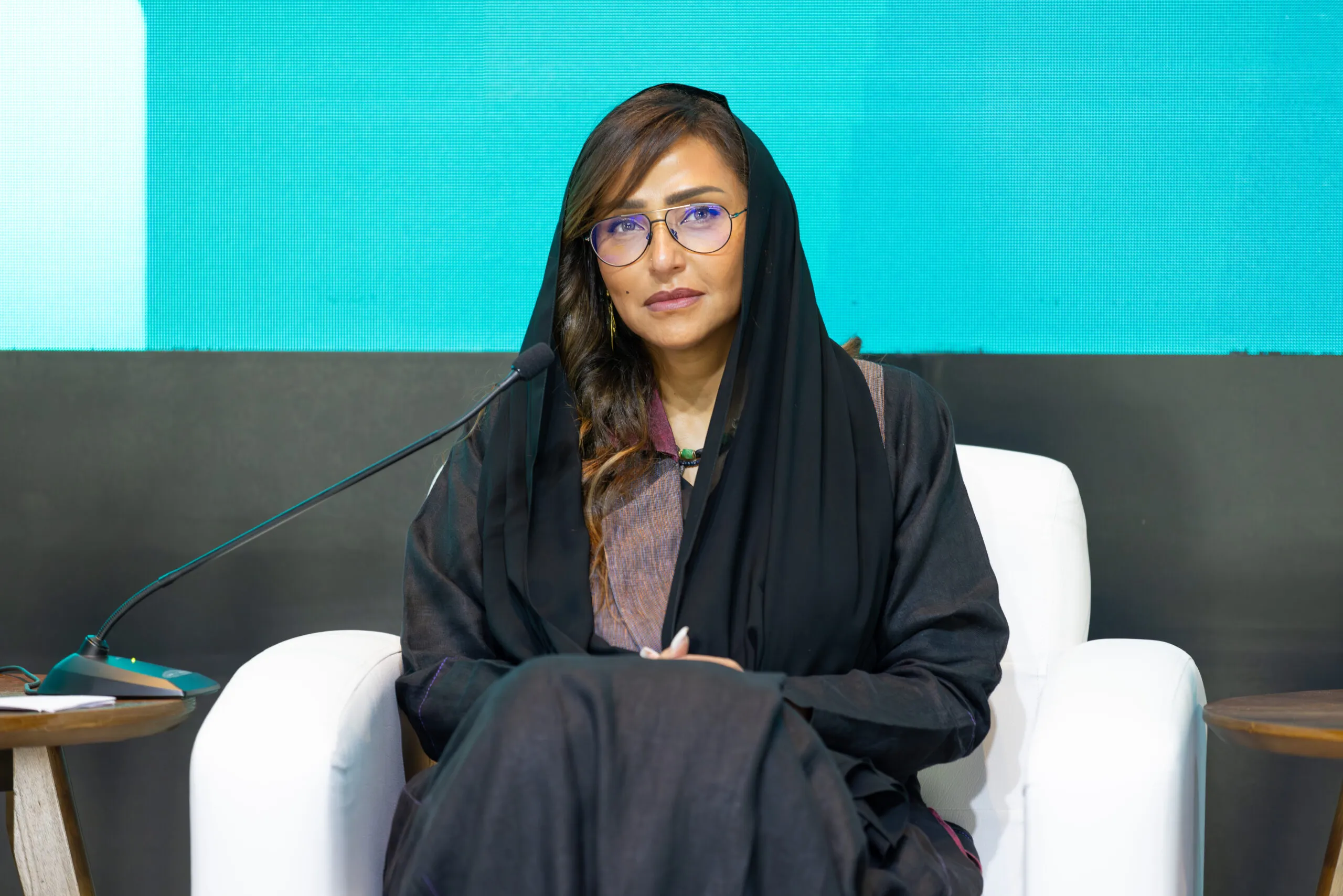 A woman seated in a white armchair before a teal backdrop, waiting to speak