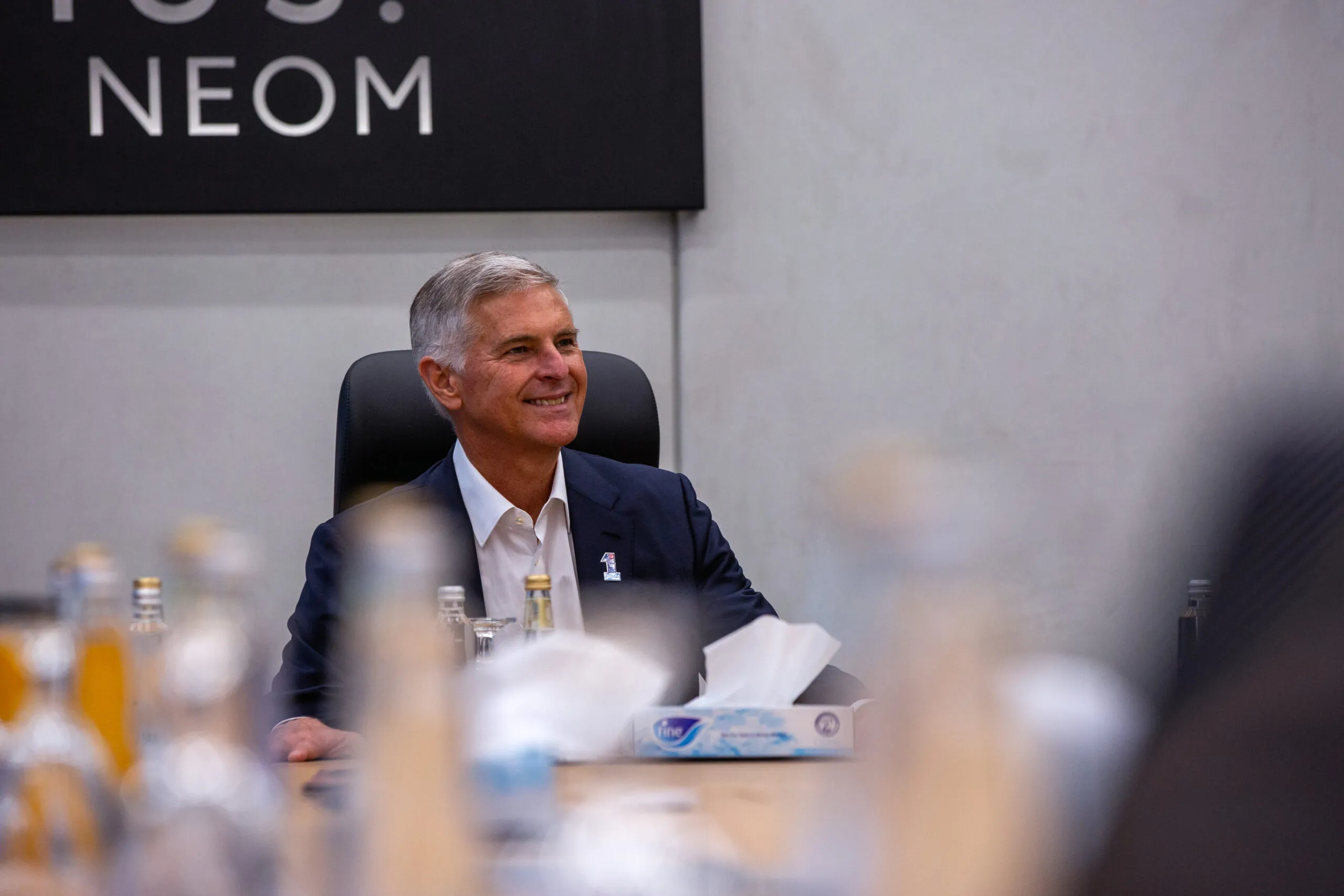 A smiling executive seated at the boardroom table beneath a NEOM sign