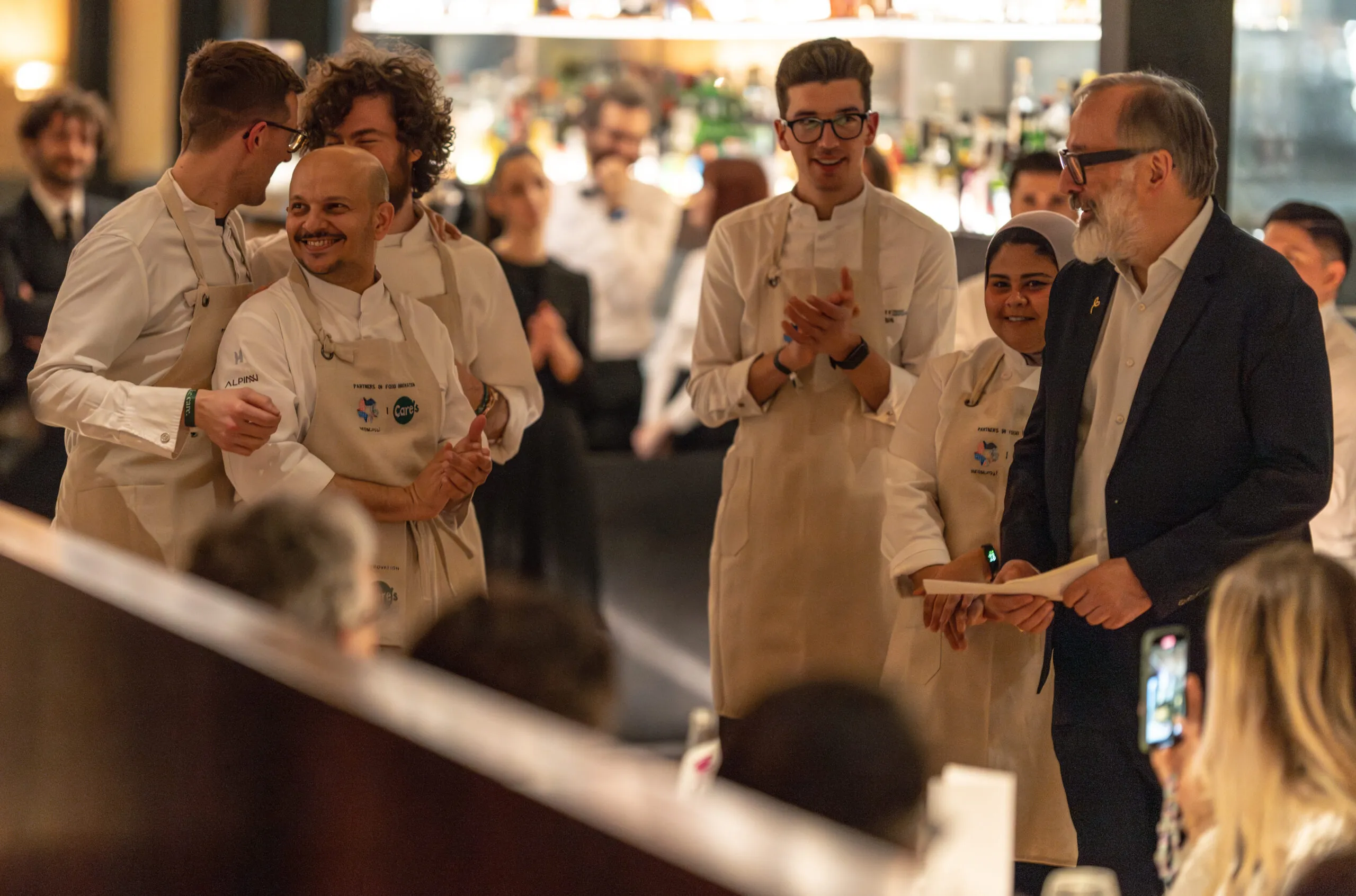 Chefs in branded aprons applauded by the dinner guests