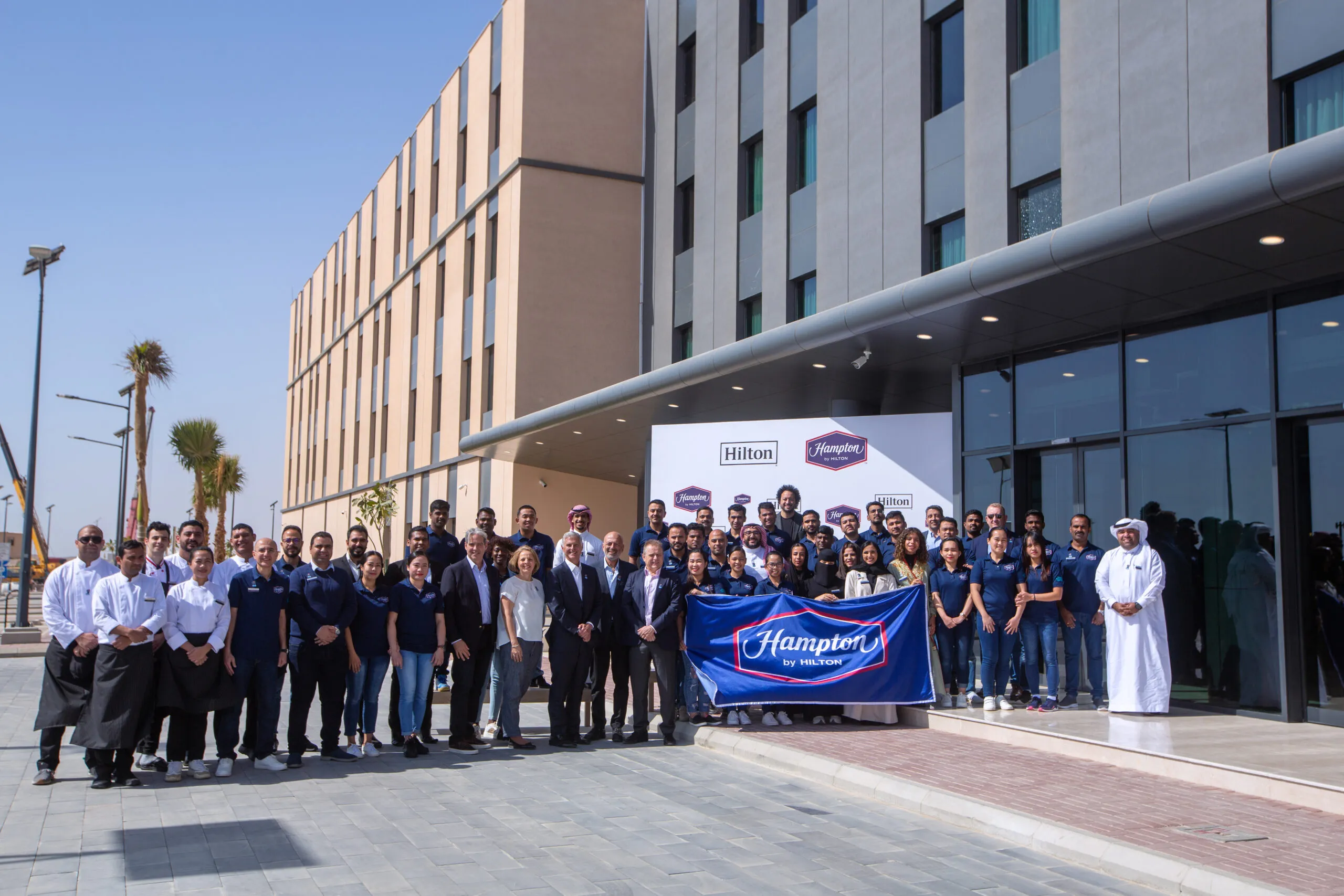 Hotel staff and leadership gathered outside the entrance with a Hampton by Hilton banner