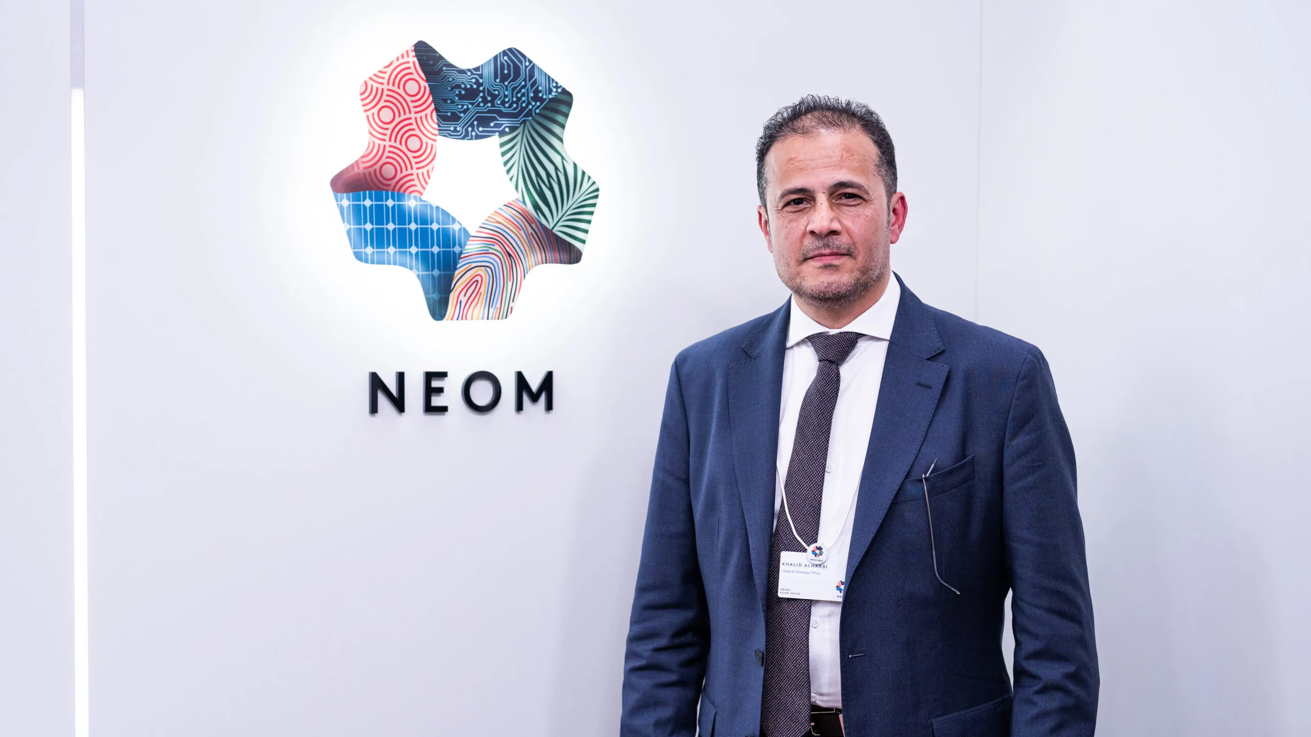 A man in a navy suit standing beneath the NEOM logo