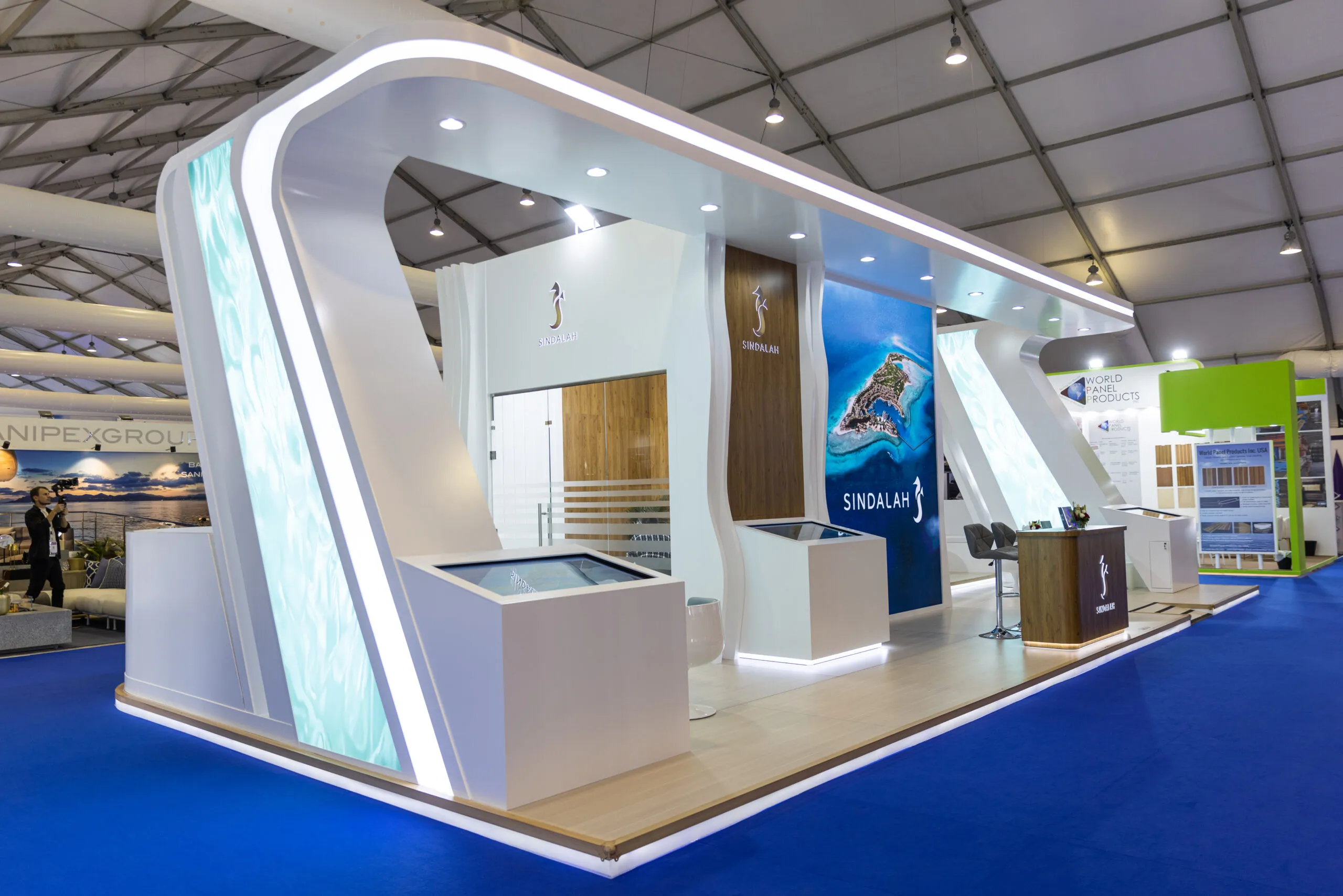 The Sindalah stand at the show — a sweeping white illuminated structure with wood panelling, standing on blue carpet inside the exhibition tent