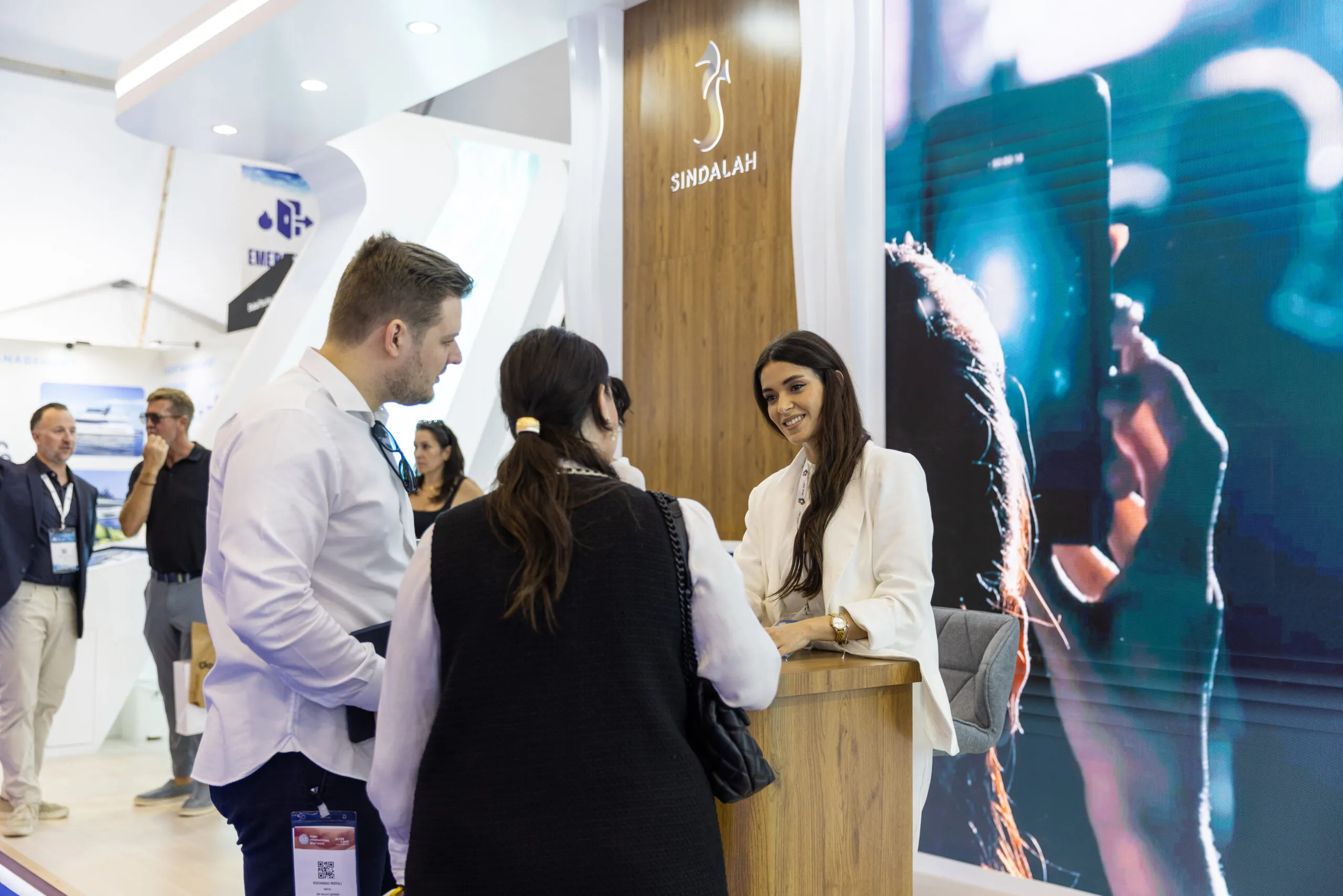 Visitors talking with staff at the reception desk of the Sindalah stand