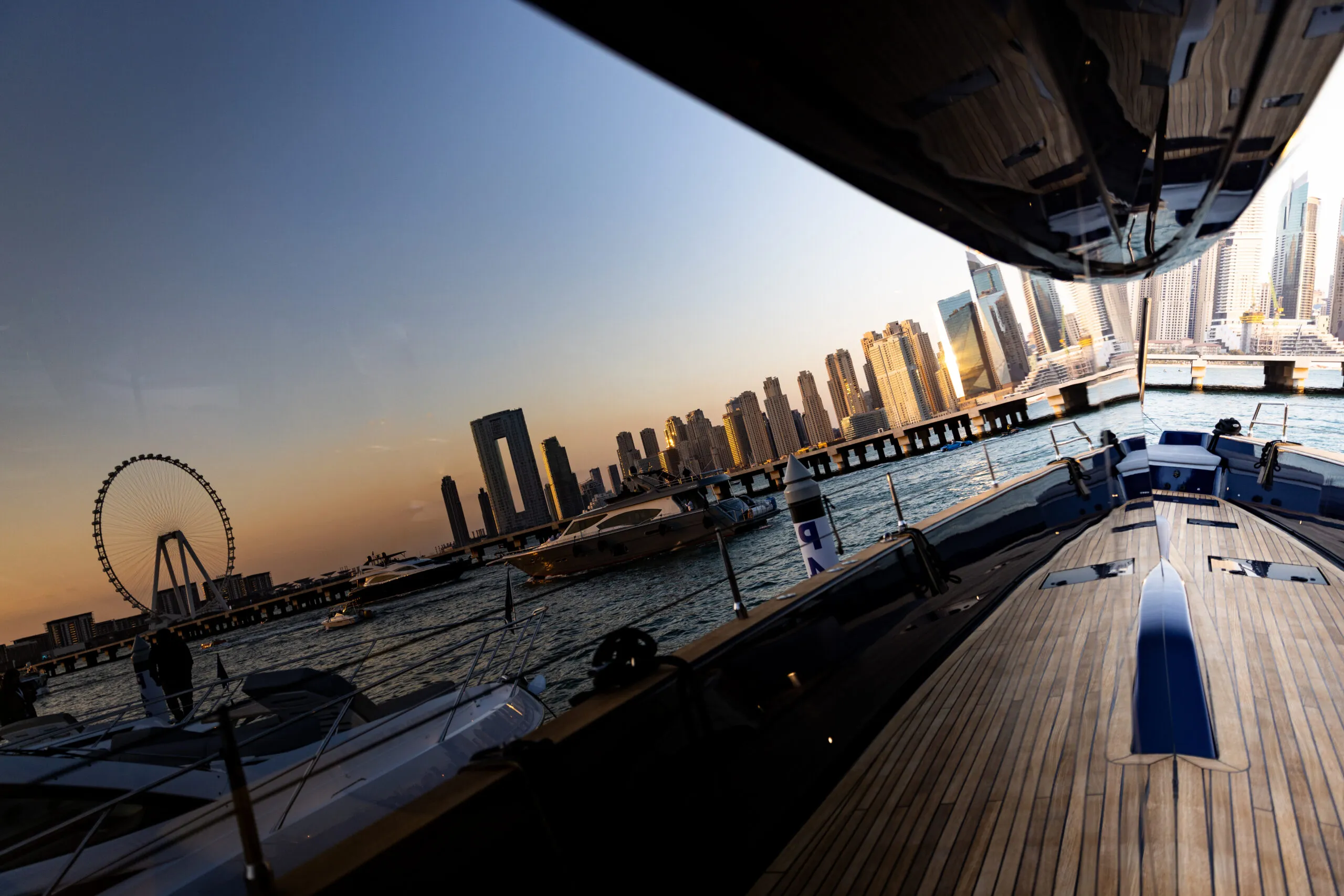 Sunset over the water from a yacht's teak foredeck, with Ain Dubai and the Dubai skyline on the horizon