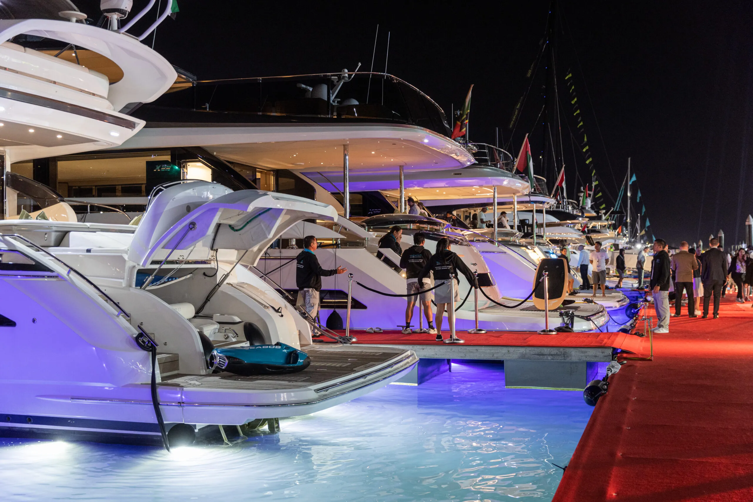 A row of illuminated motor yachts along the red-carpeted dock at night