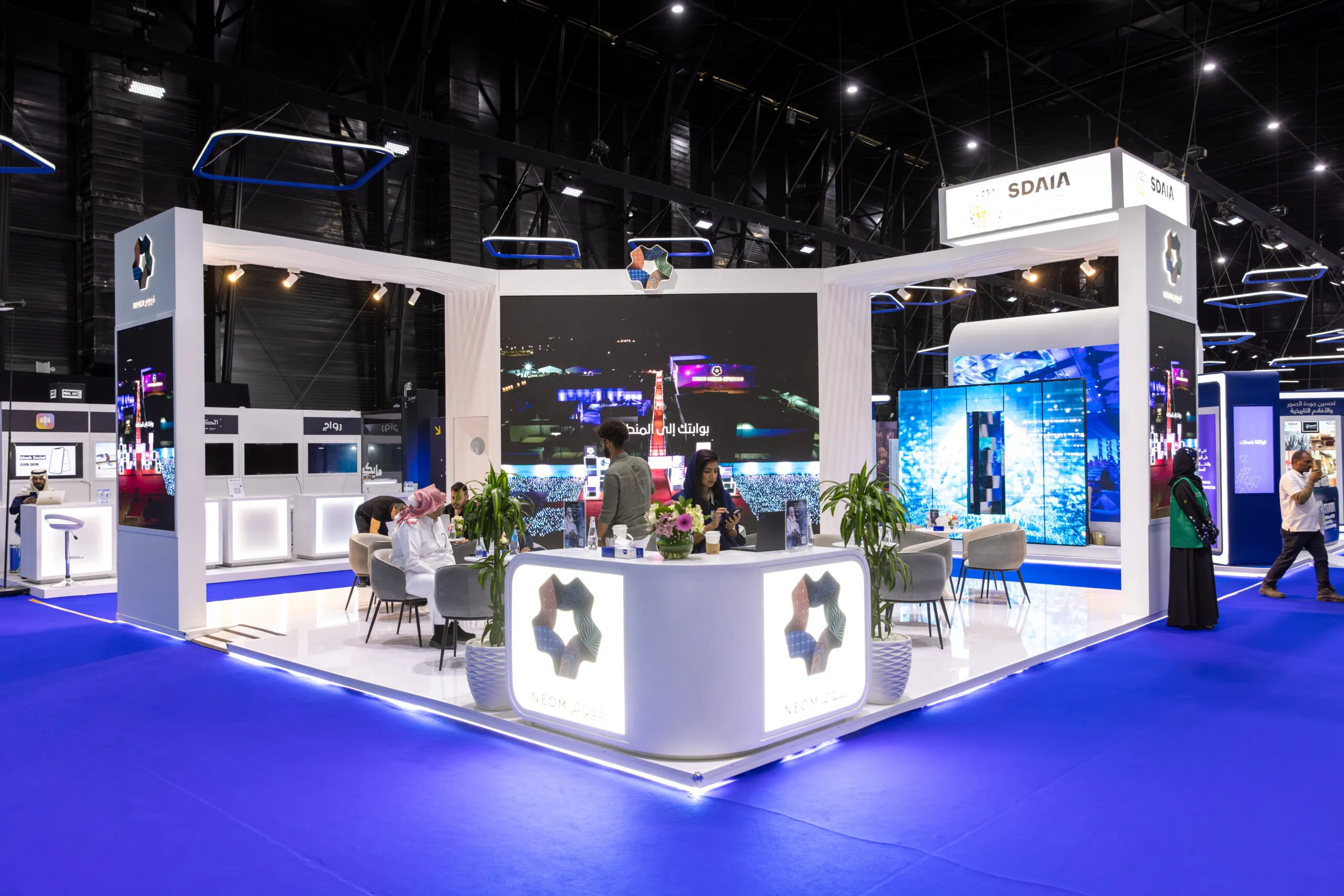 The NEOM stand on the blue exhibition floor, with lounge seating and video walls