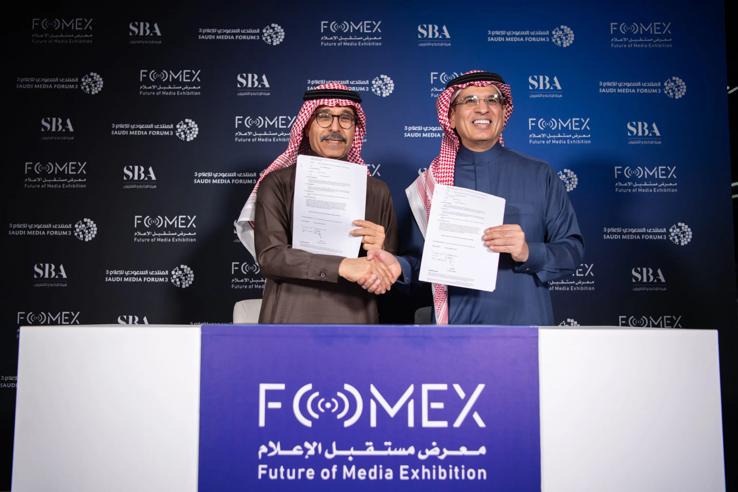 Two men holding up signed agreements at the FOMEX — Future of Media Exhibition — signing desk
